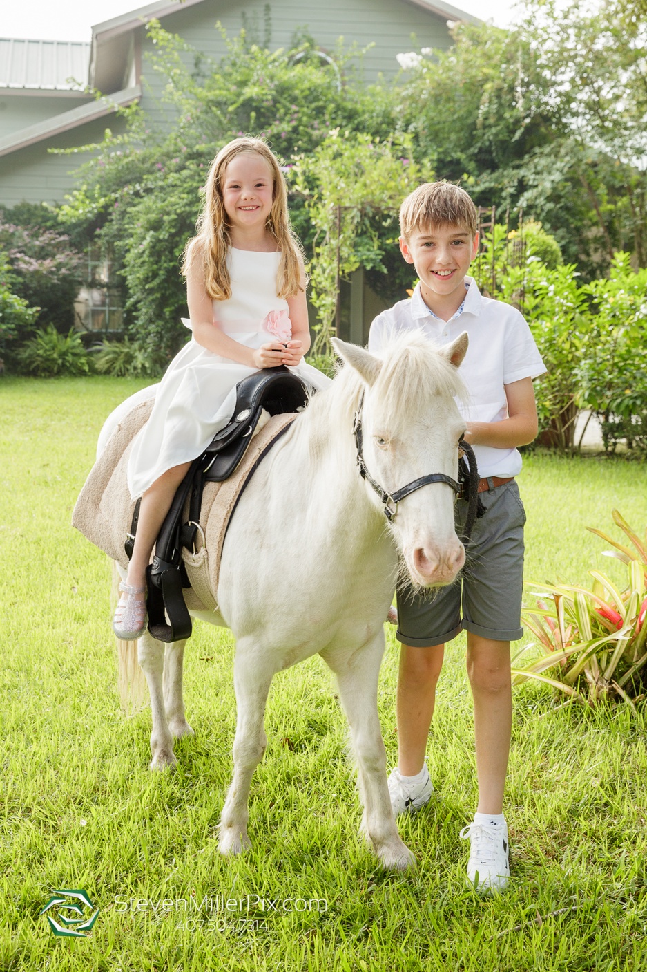 The little girl sitting on the small white horse Bandit while her brother stands beside her as they stand in the grass and pose of a picture