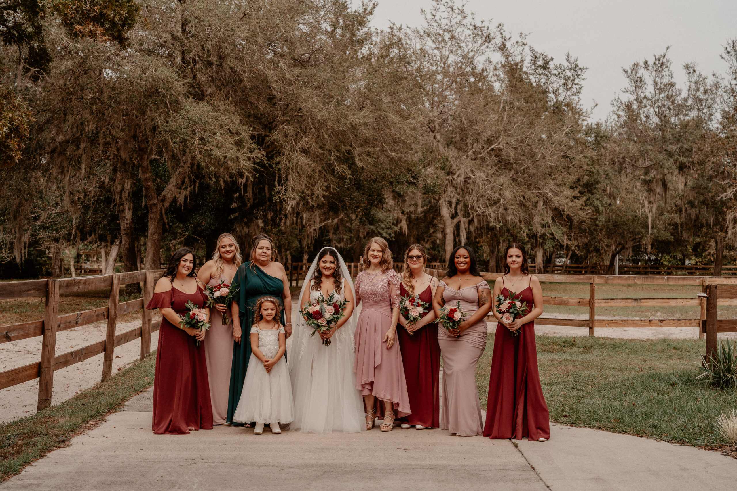 Bridesmaids in maaroon with bride and little girl in front of a fence and field