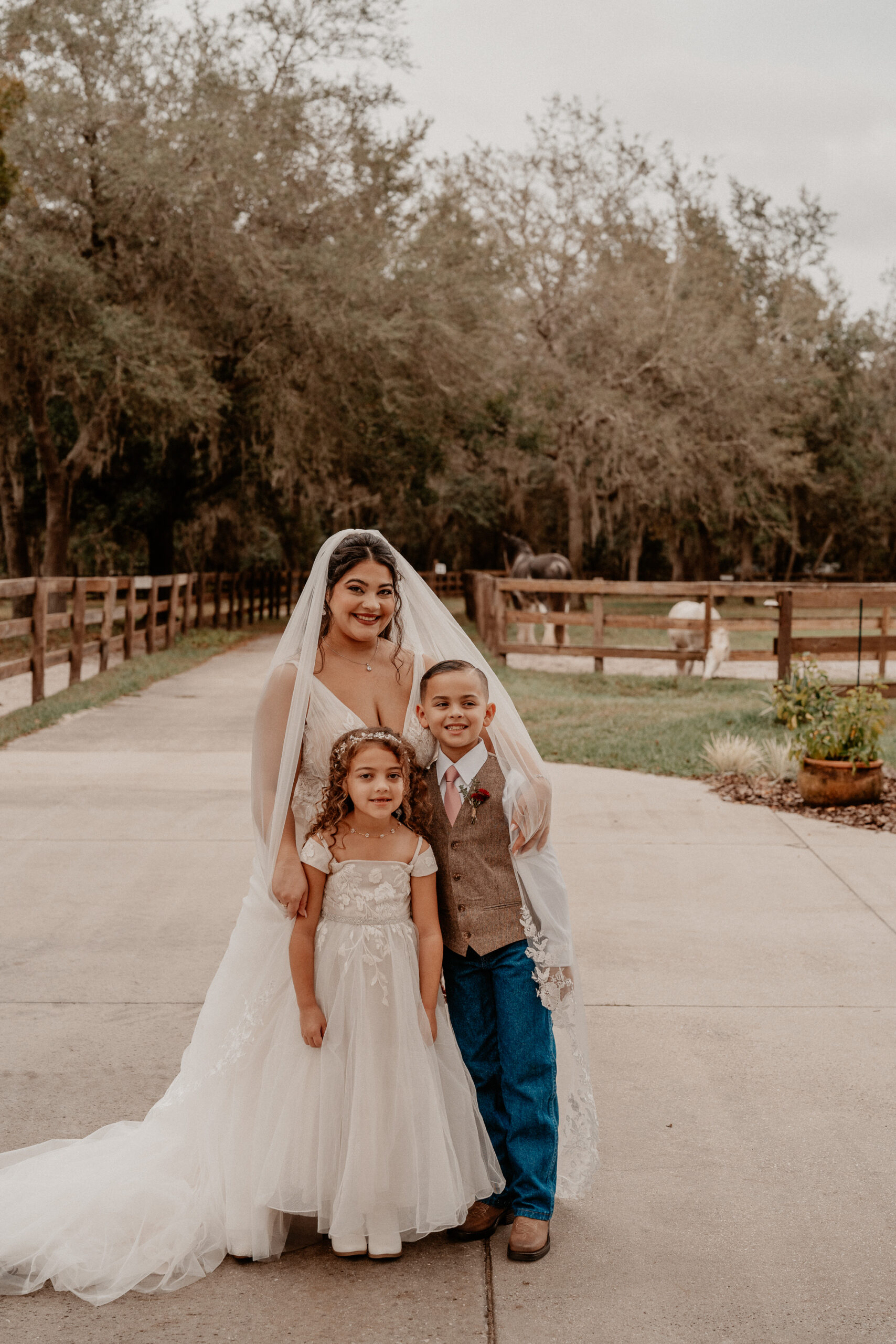 Bride with a little girl and a little boy in front of a fence with horses