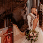 The happy couple kissing in the barn with a beautiful black horse looking upon them at BLB Hacienda